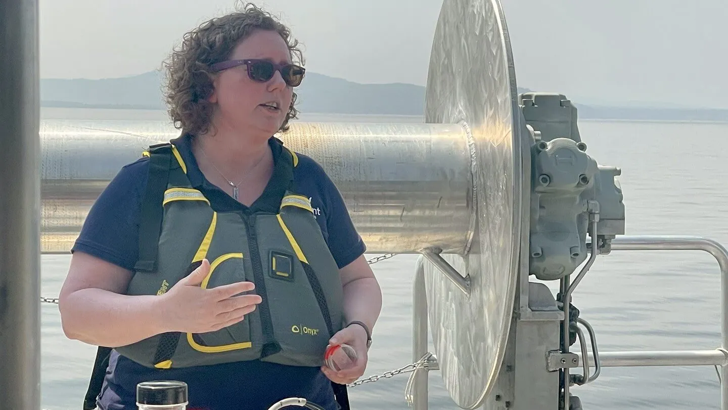 Anne Jefferson stands on a boat and discusses her research. Anne is wearing sunglasses, a tee shirt and a life or flotation vest. A lake and mountain are in the background.