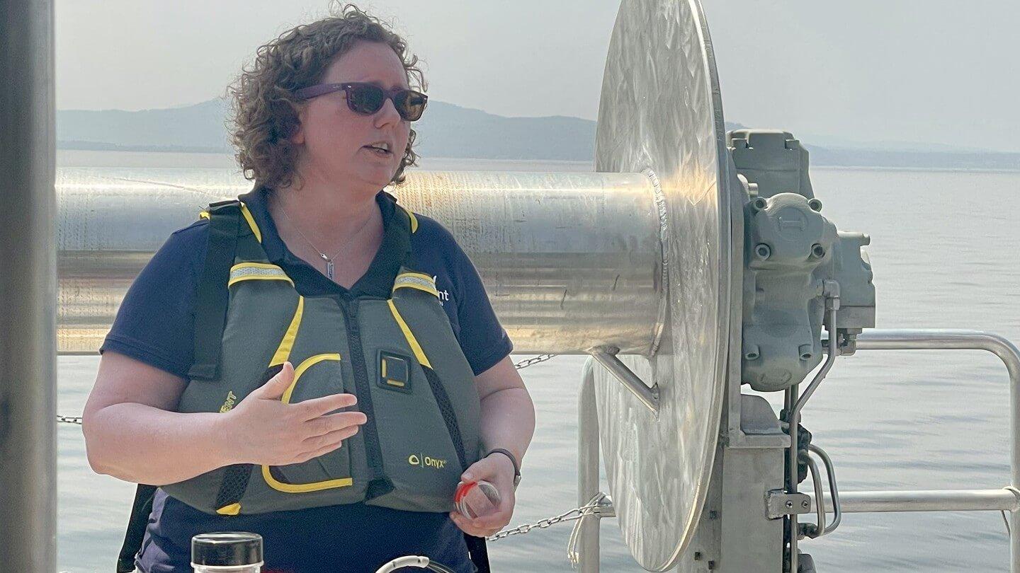 Anne Jefferson stands on a boat and discusses her research. Anne is wearing sunglasses, a tee shirt and a life or flotation vest. A lake and mountain are in the background.