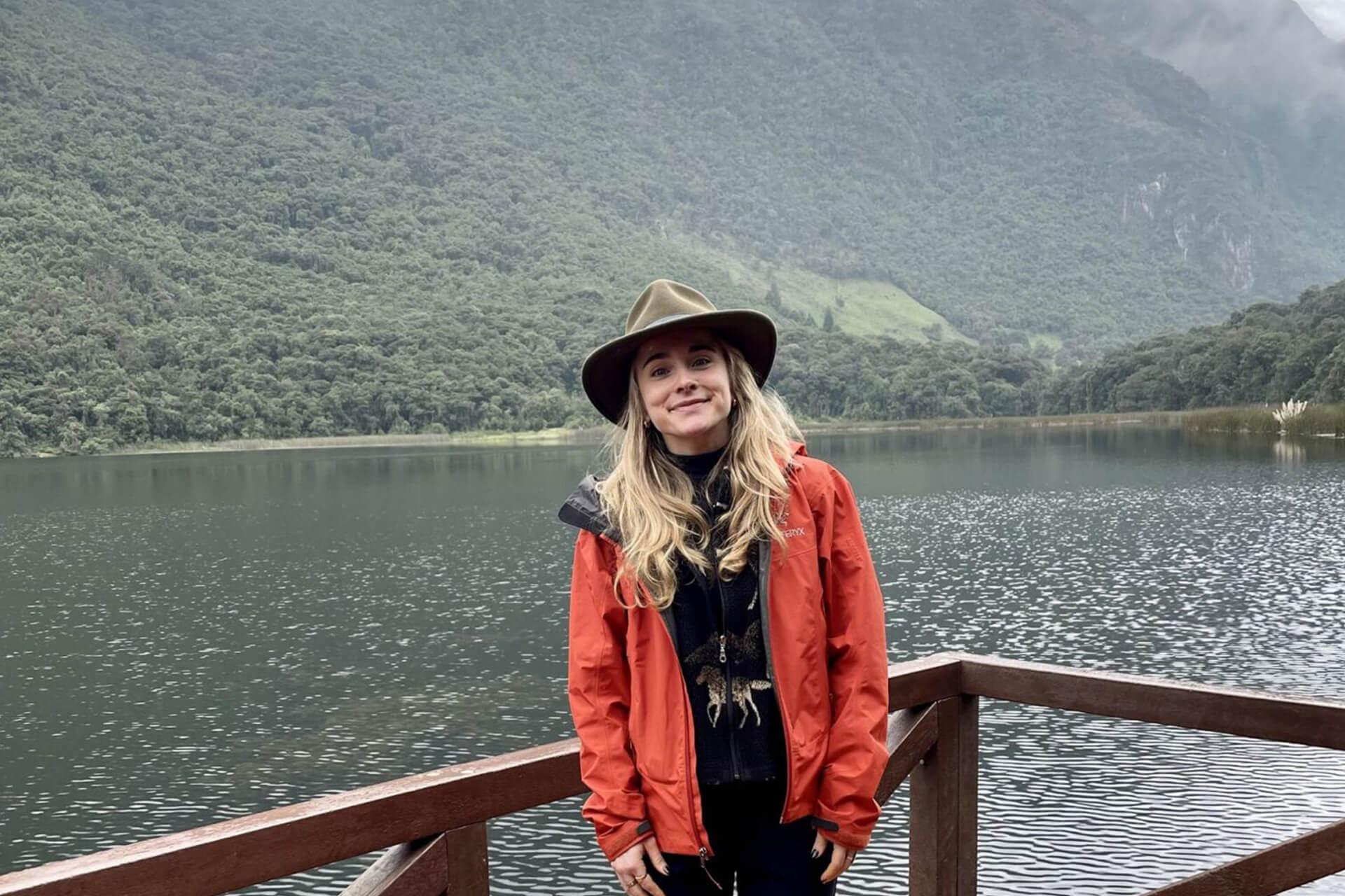 A smiling woman wearing a hat and a red jacket stands on a wooden deck in front of a calm lake and a green, tree-covered mountain under a cloudy sky. 