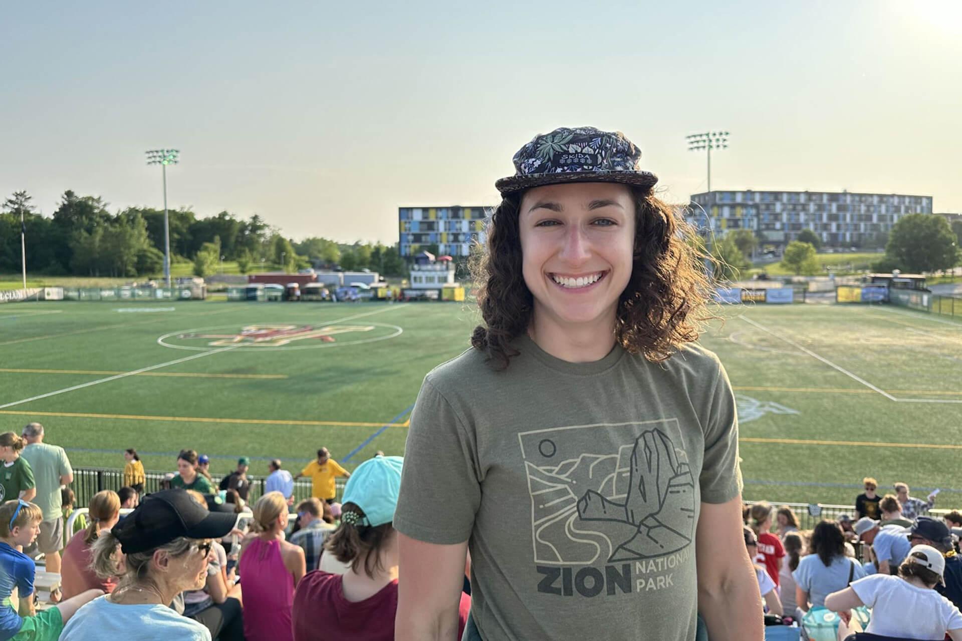 A person wearing a patterned hat and a t-shirt with a Zion National Park logo smiles in a stadium. The background shows a crowd in the stands and a sports field.