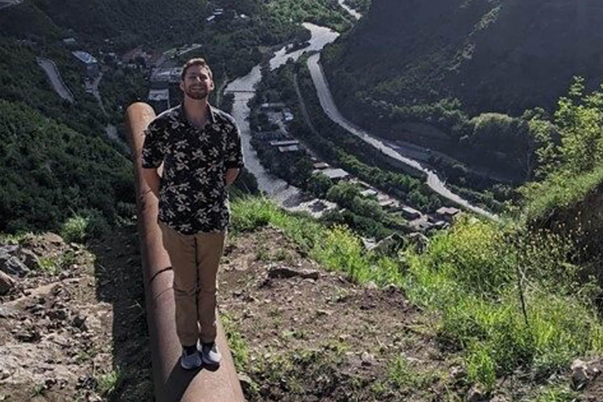 A man in a floral shirt and tan pants stands on a large pipe on a hillside, with a winding river and a road visible in a deep valley below.