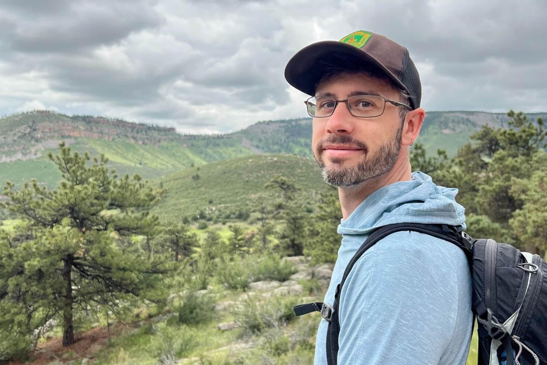 A man with glasses and a beard, wearing a baseball cap and backpack, stands outdoors with green hills and trees in the background under a cloudy sky.