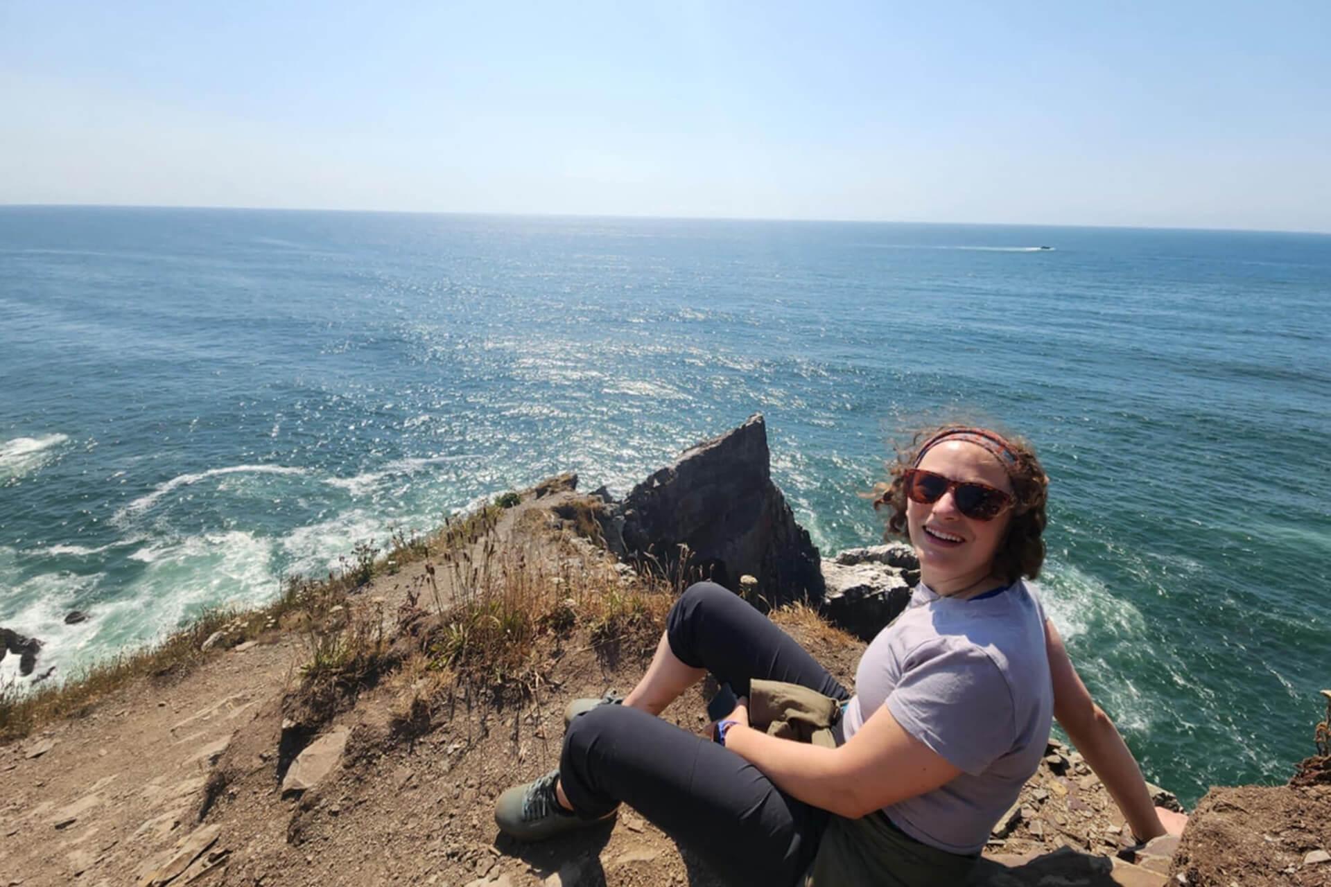 A smiling woman sits on a rocky cliff overlooking a blue ocean.