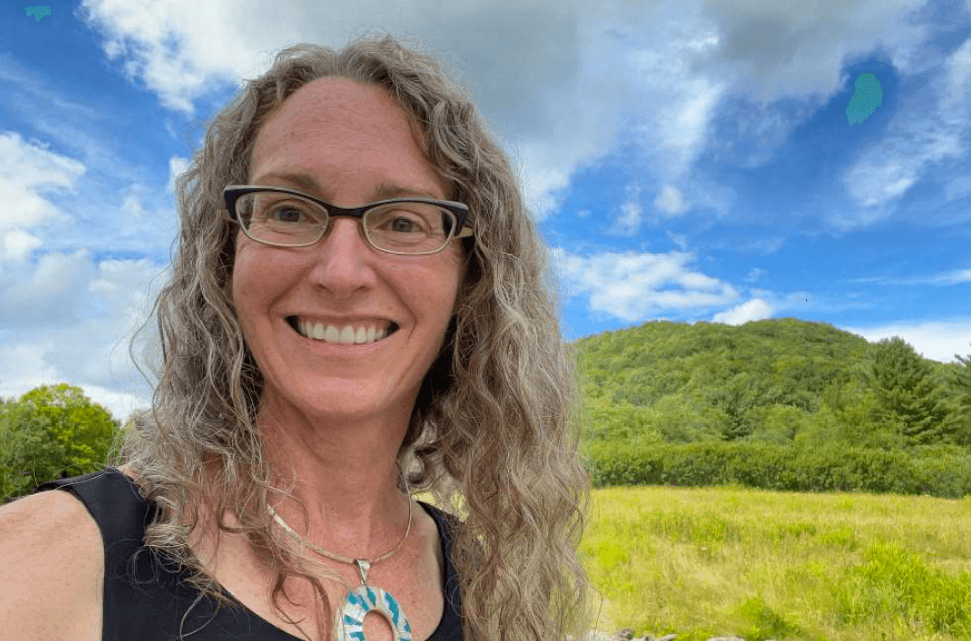 A woman wearing glasses smiles. Green grass and blue sky are behind her.