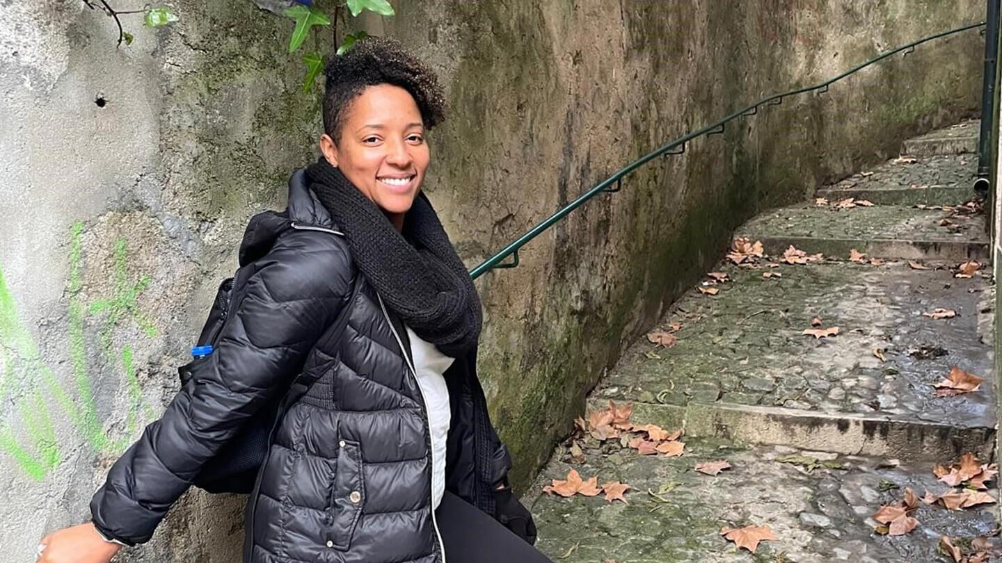 A person is smiling at the camera, wearing a black puffy jacket, a black knitted scarf, and a backpack.