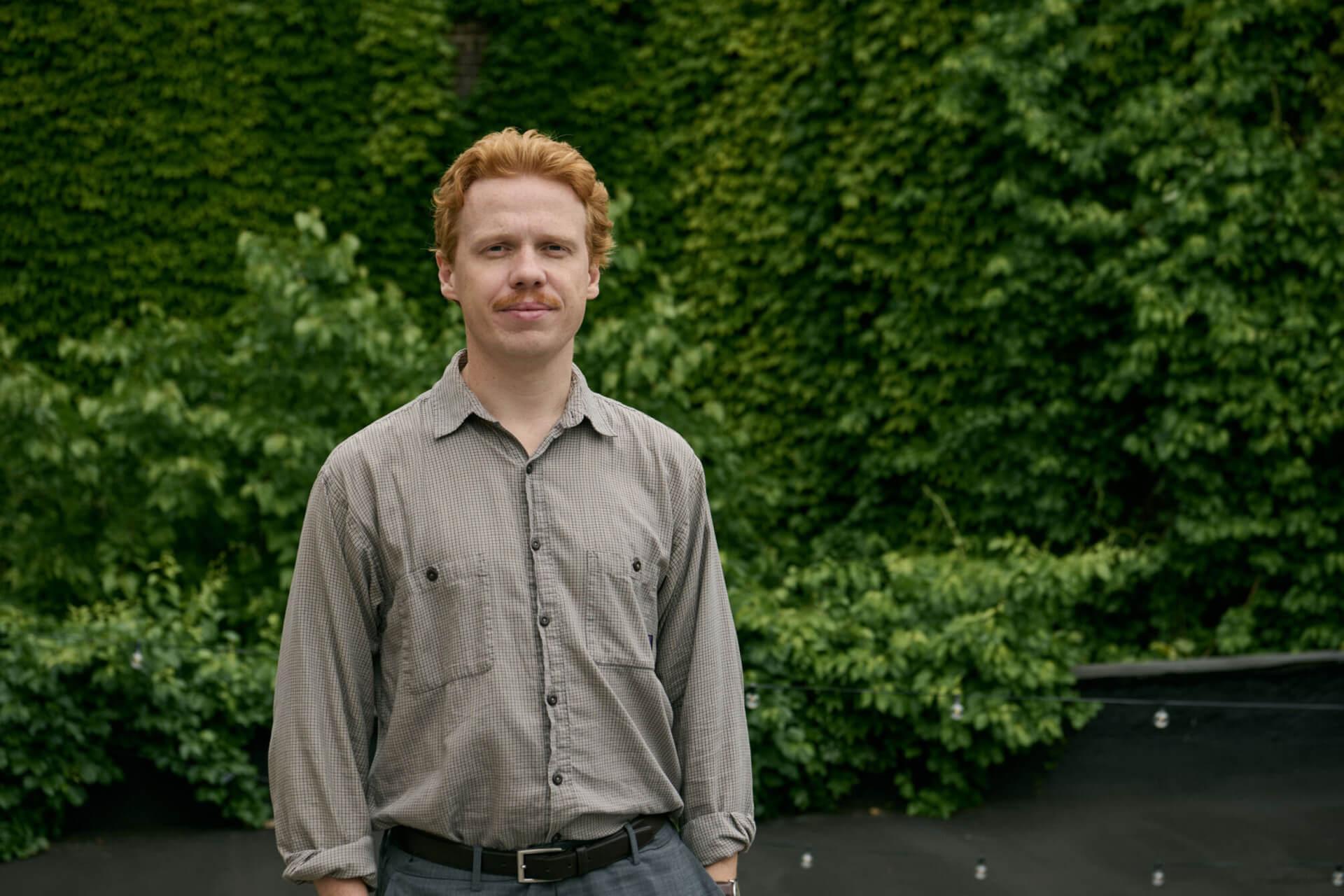 A man is standing in front of a wall of green ivy. 