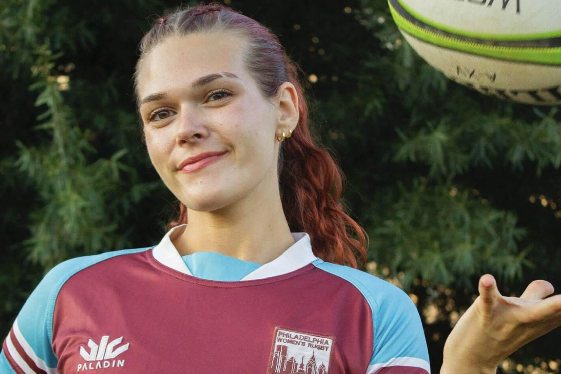 A person in a red and blue Philadelphia Women's Rugby jersey is smiling and looking at the camera, with a rugby ball hovering in the air to their right.