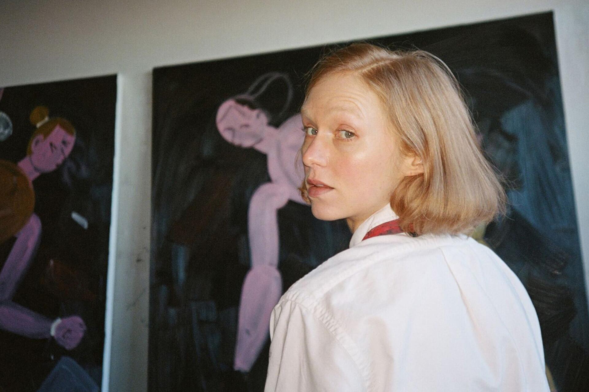 A person with blonde hair wearing a white shirt looks over their shoulder at the camera, with two paintings visible in the background.
