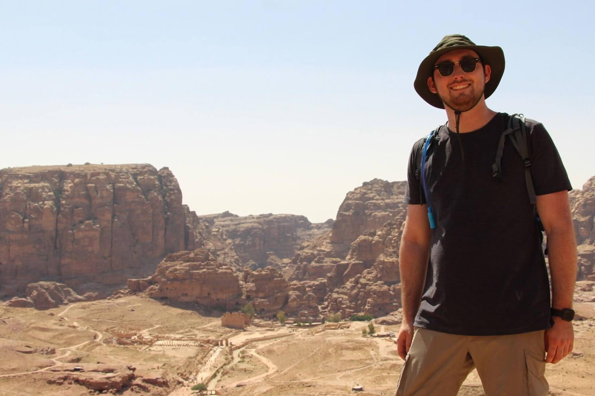 A person wearing a sun hat and sunglasses, with a backpack, stands on a rocky overlook, smiling at the camera. In the background is a vast, arid canyon landscape with a path winding through it.