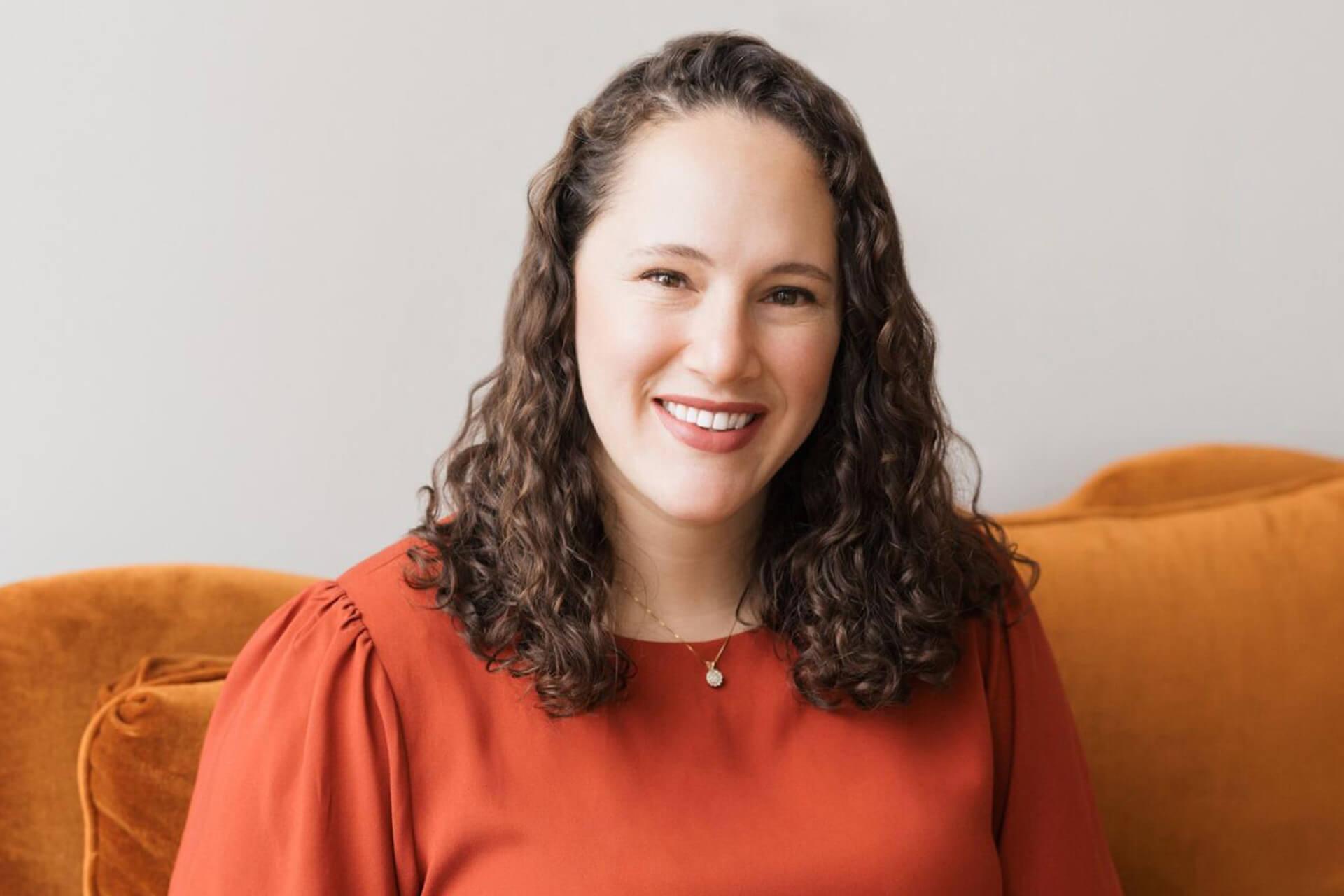 A smiling woman sits on an orange couch indoors.