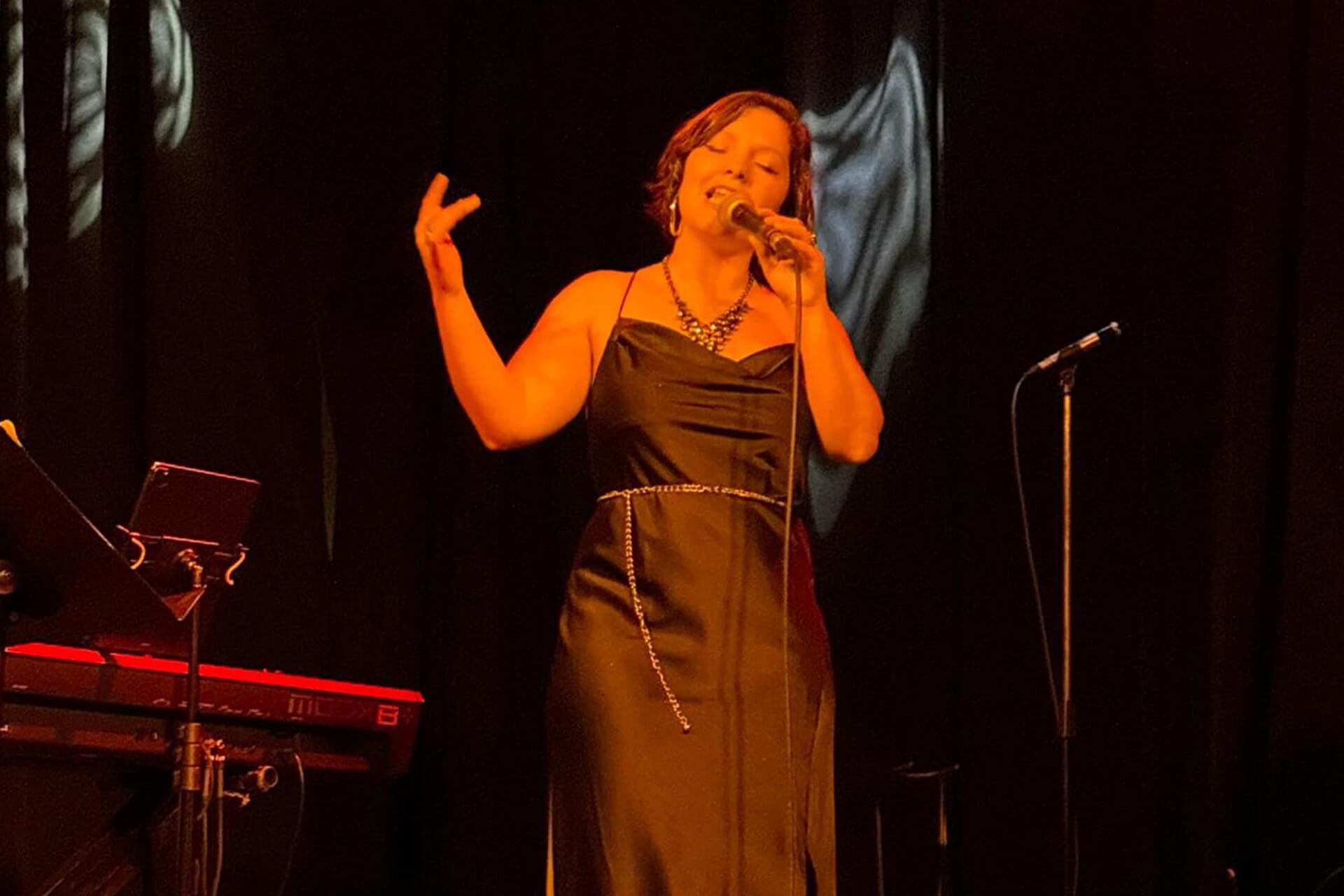 A woman in a black dress sings into a microphone on a stage, with a keyboard and dark curtains in the background.