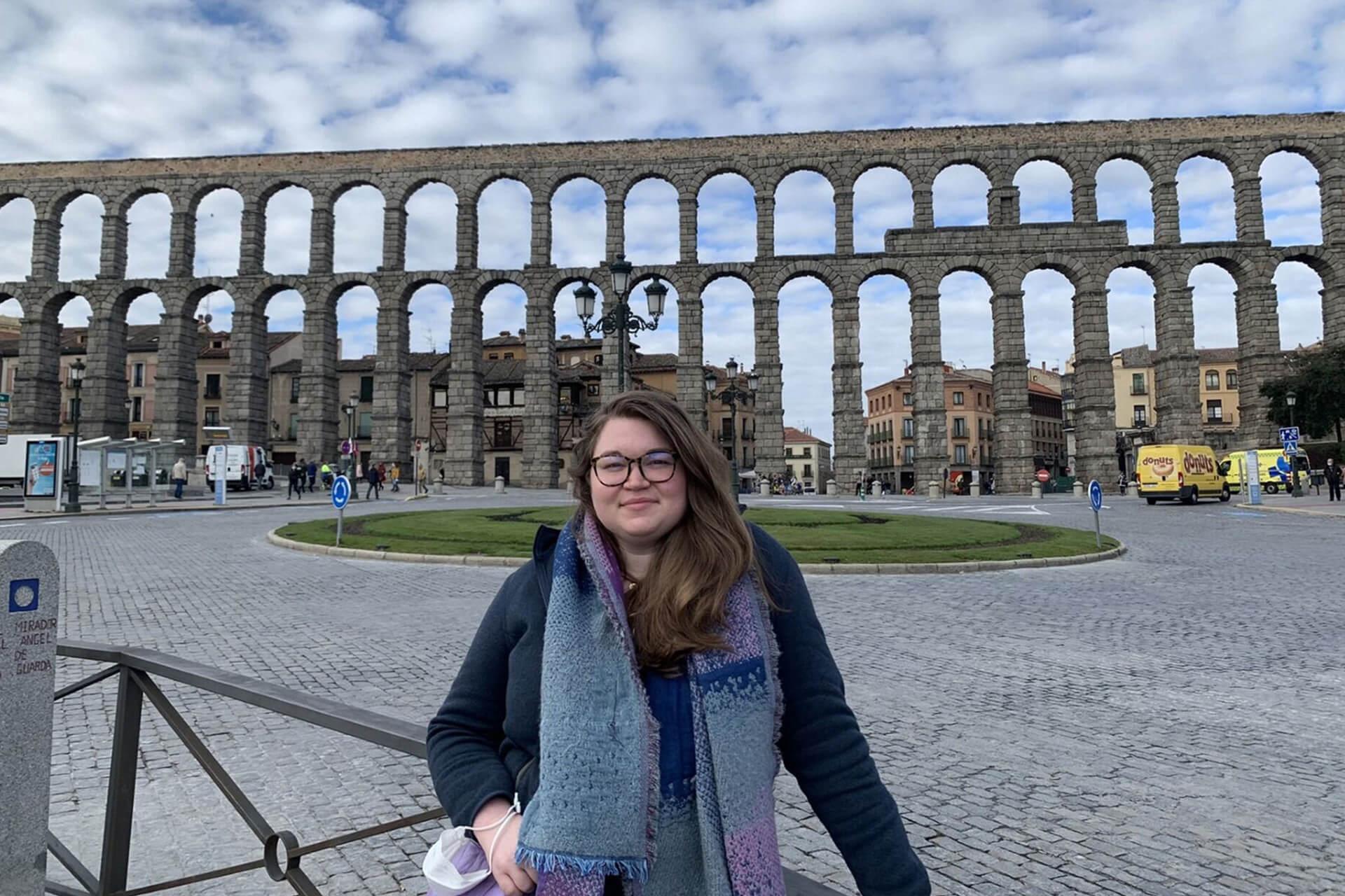 A woman stands in front of a large ancient Roman aqueduct, wearing a coat and a long scarf.