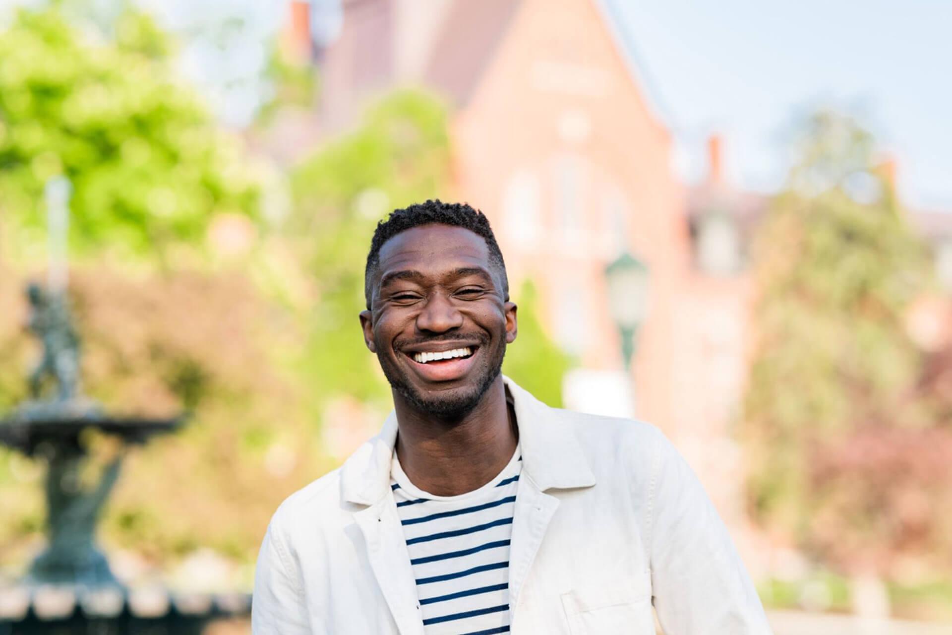 A man smiles brightly at the camera, standing in a park with a fountain and a brick building in the background.          Analysis