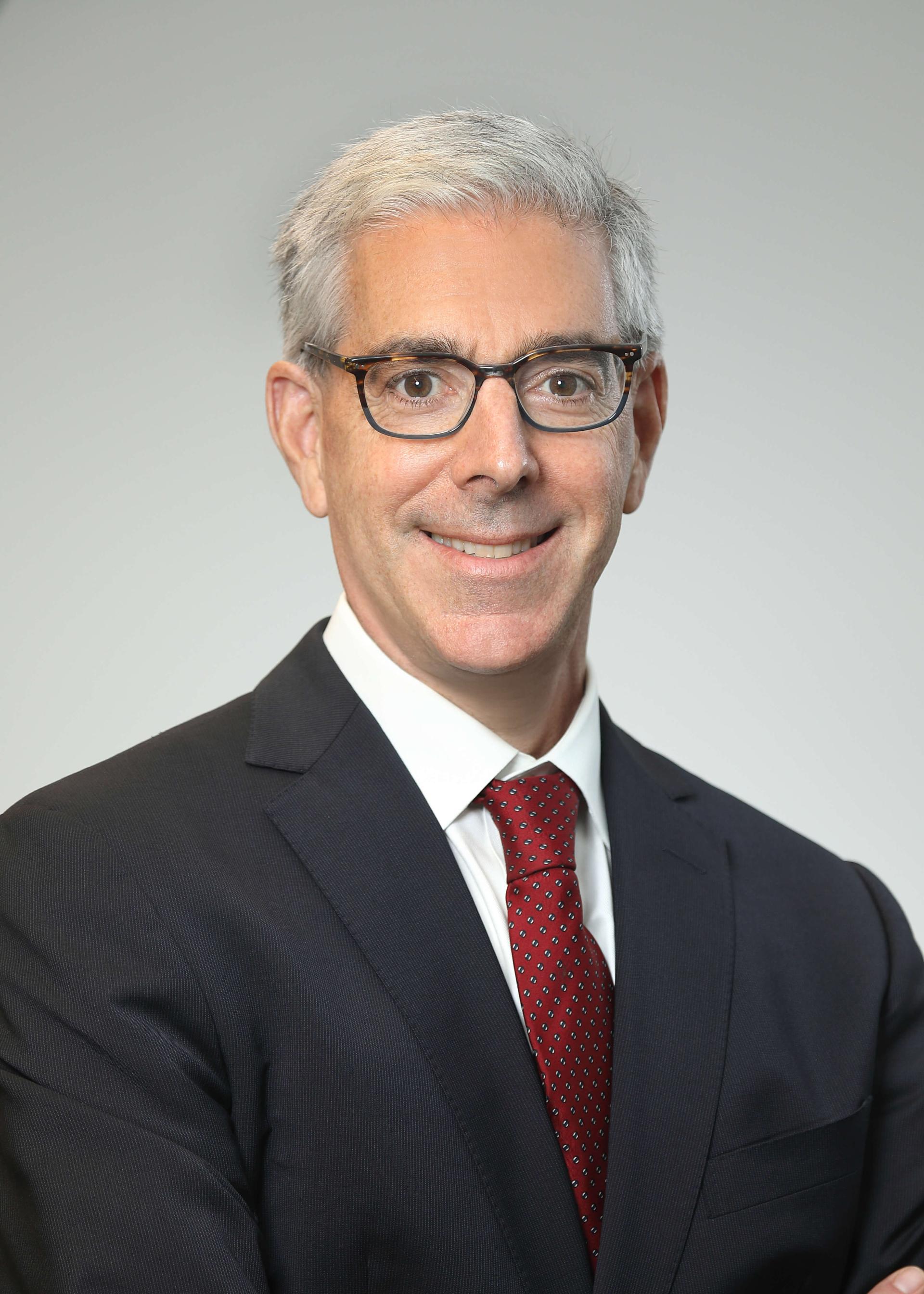 Man in dark suit, red tie and white shirt with glasses poses for a headshot.