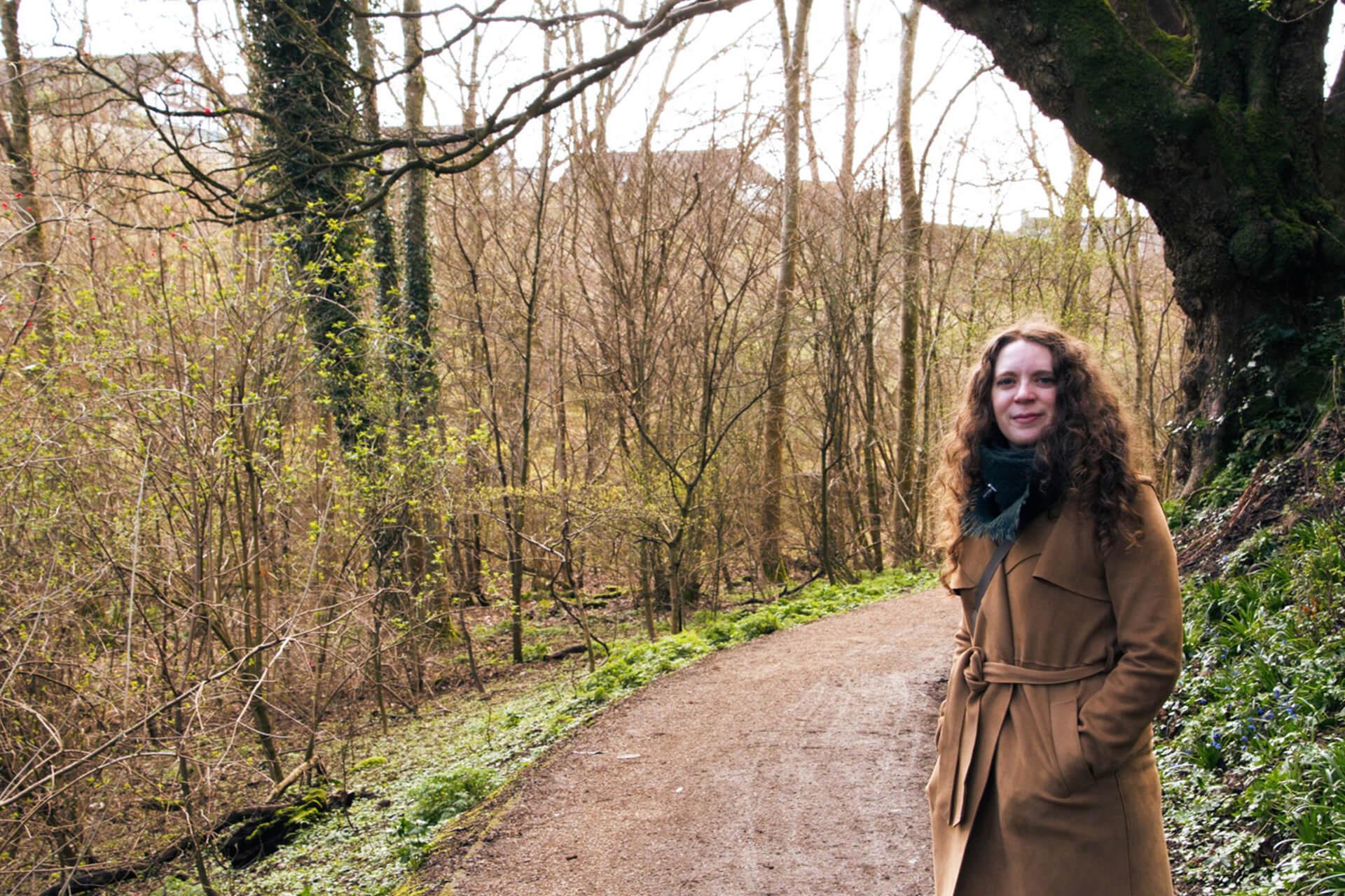A woman wearing a tan trench coat stands on a dirt path in a wooded area. She is looking at the camera with a slight smile.