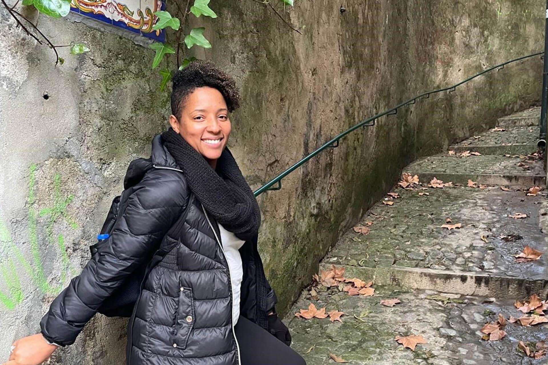 A person in a black puffer jacket and scarf smiles while standing on a cobblestone path next to a stone wall with a handrail and fallen leaves.