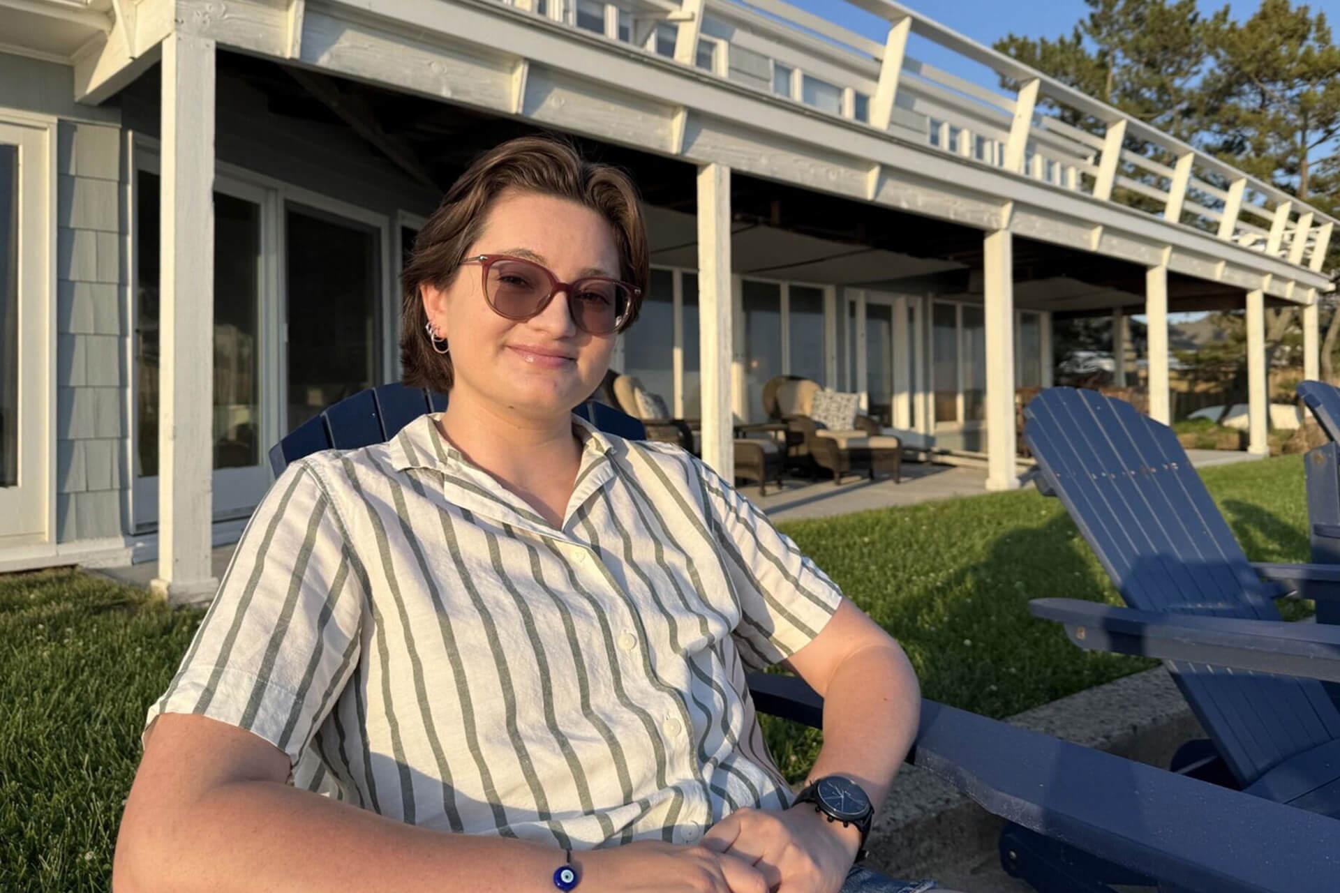 A person with sunglasses sits in a blue Adirondack chair outdoors, smiling in front of a house with a porch and a grassy lawn. 