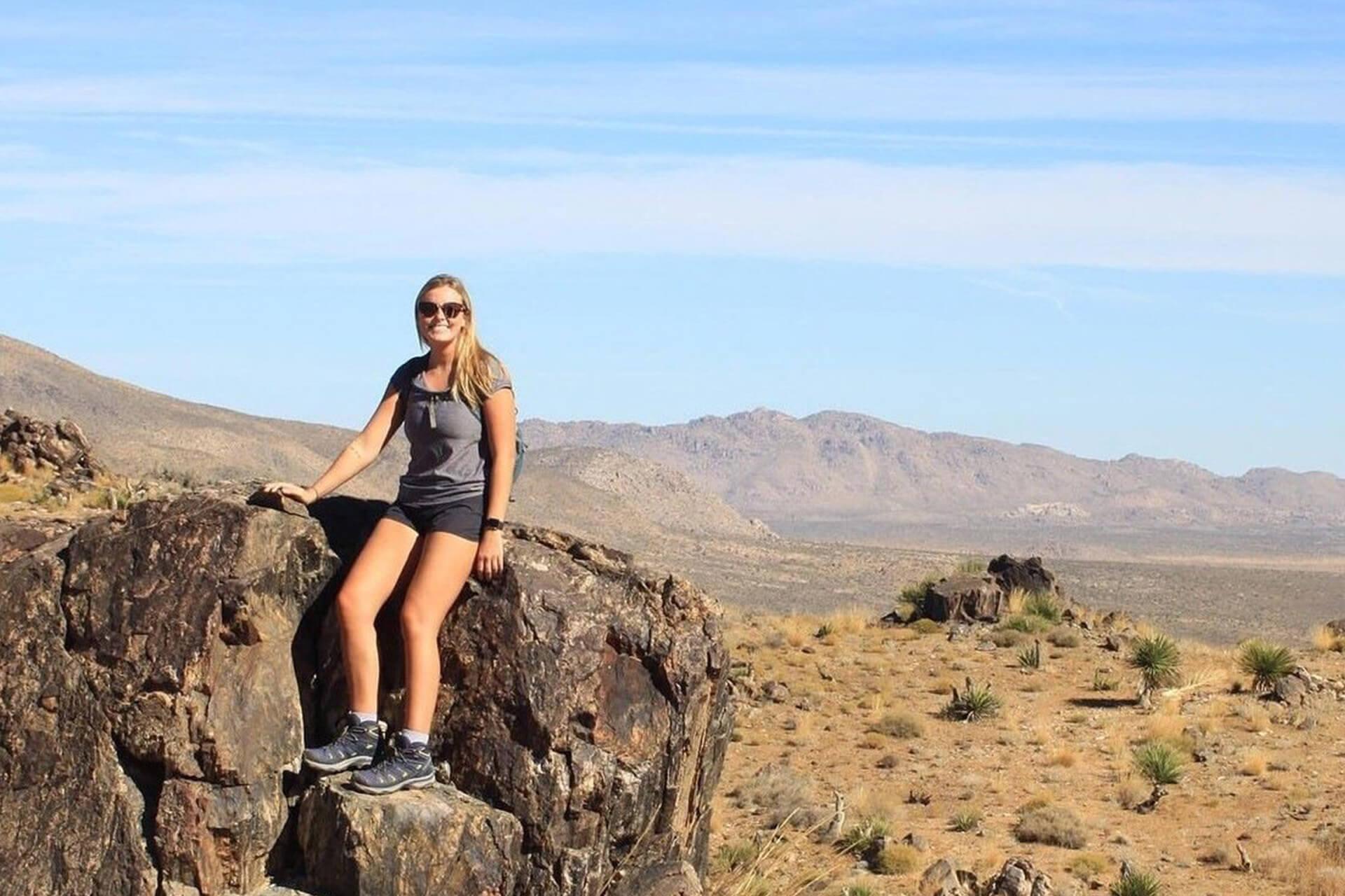 A smiling woman in sunglasses, shorts, and a t-shirt is sitting on a large rock in a desert landscape. In the background are hills and sparse vegetation under a clear blue sky.