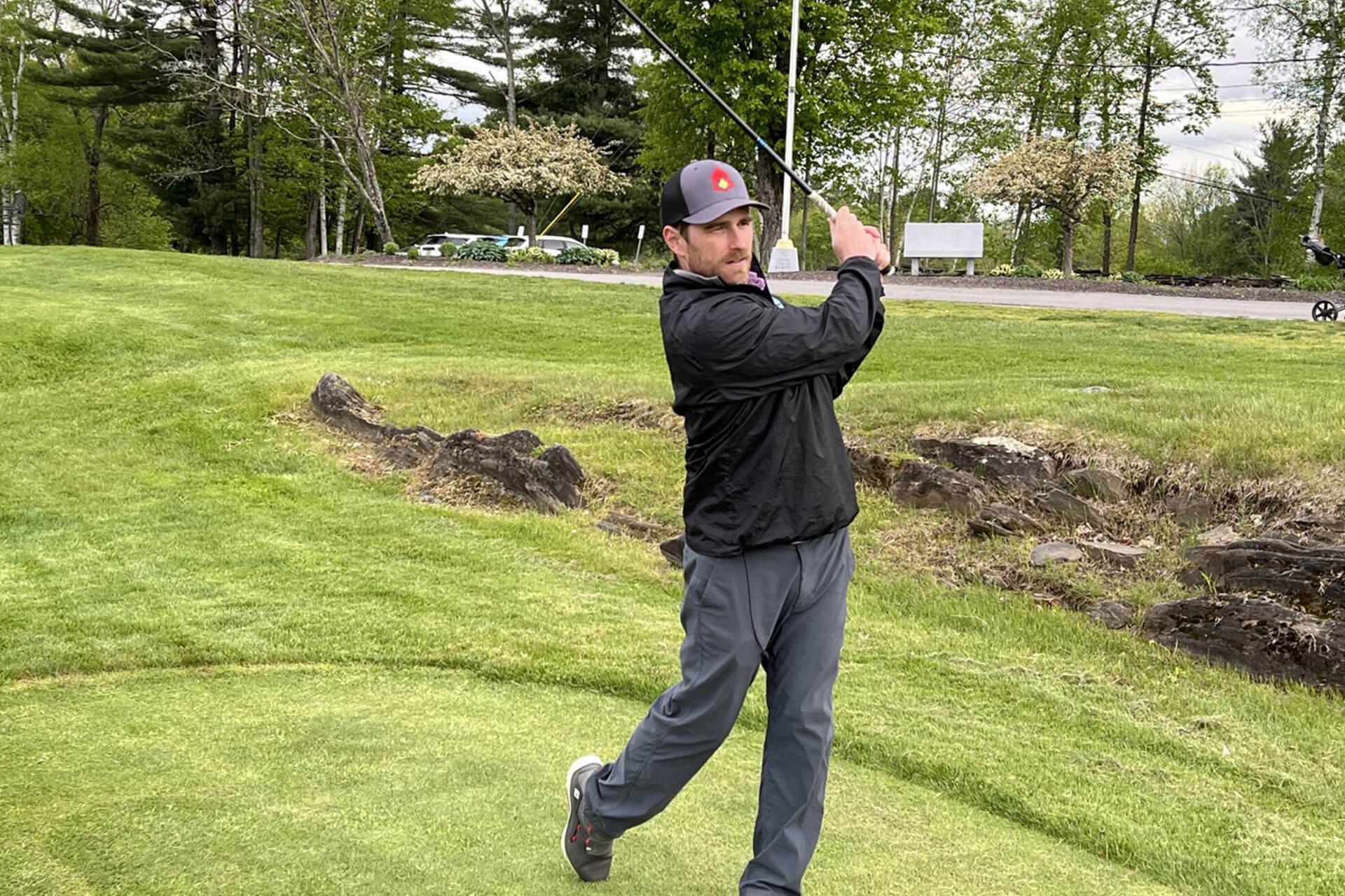 A man in a black jacket and a baseball cap swings a golf club on a green course. An American flag is in the background.