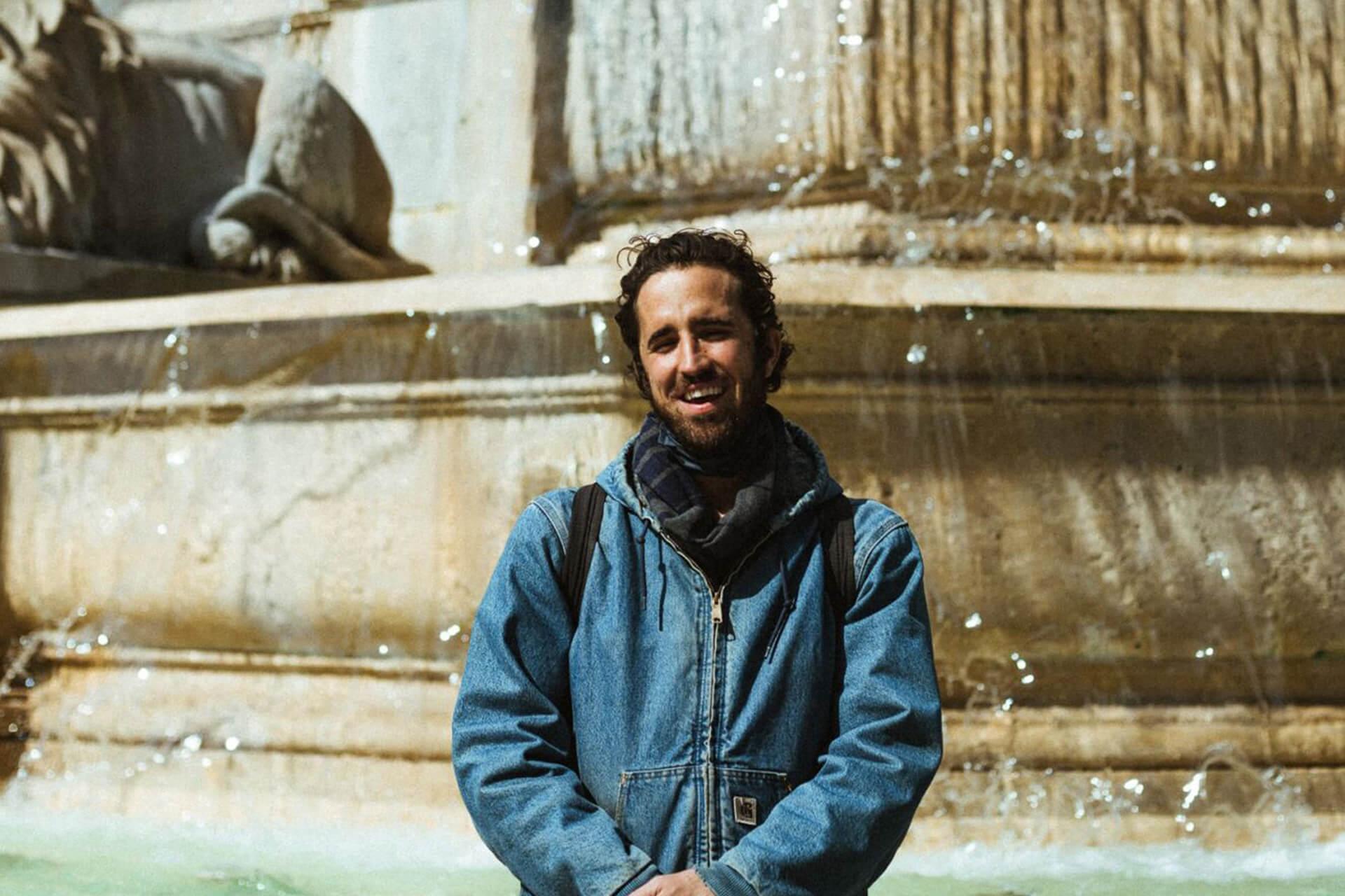 A smiling man with a beard and curly hair stands in front of a stone fountain with water splashing. He is wearing a denim jacket and a dark scarf.