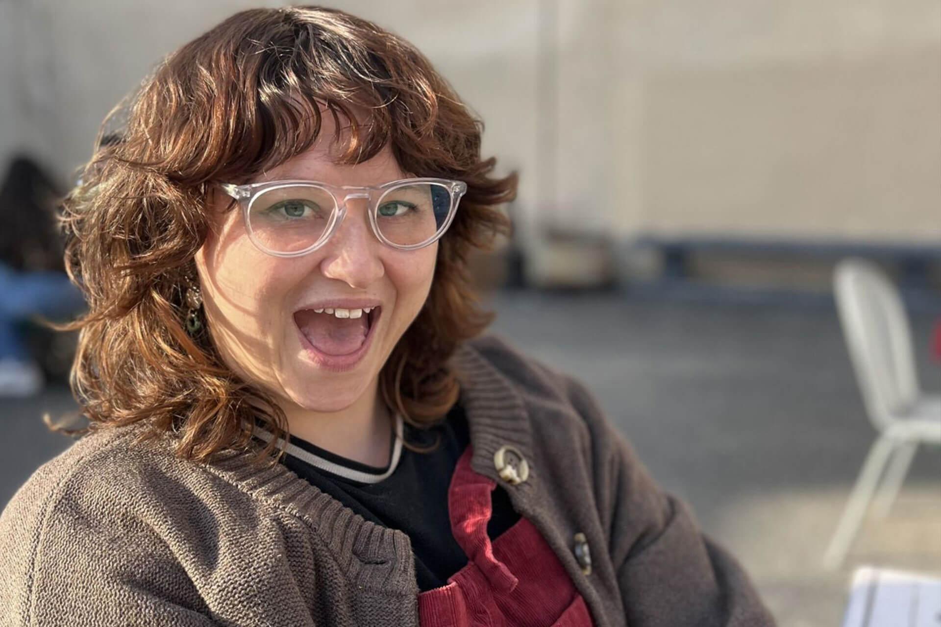 A person with glasses smiles with an open mouth, wearing a cardigan over a dark shirt and a red apron. The background is a blurry outdoor setting.