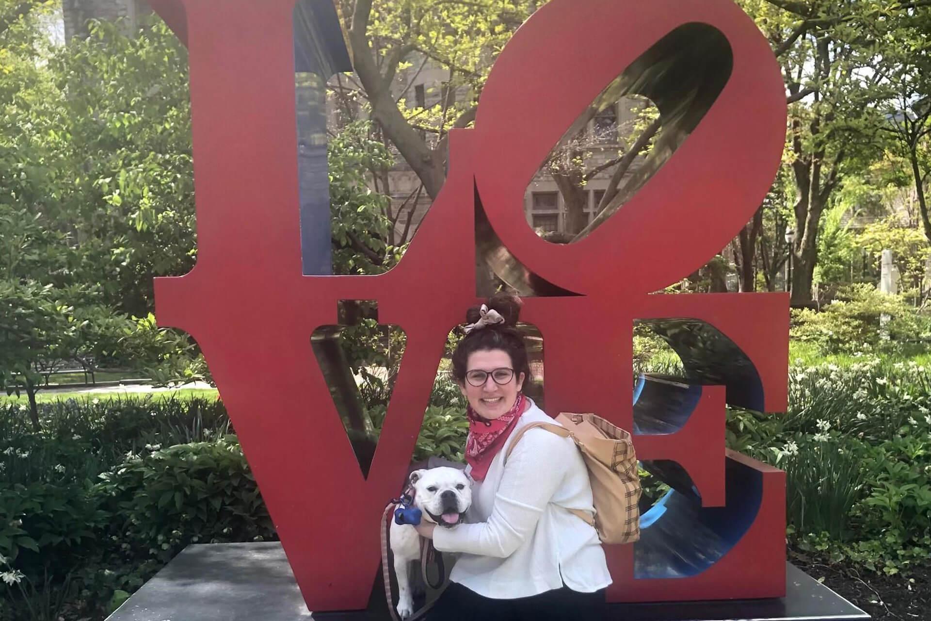 A woman holds a dog in front of a large red love sculpture