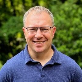 A man in a blue shirt and glasses smiles.