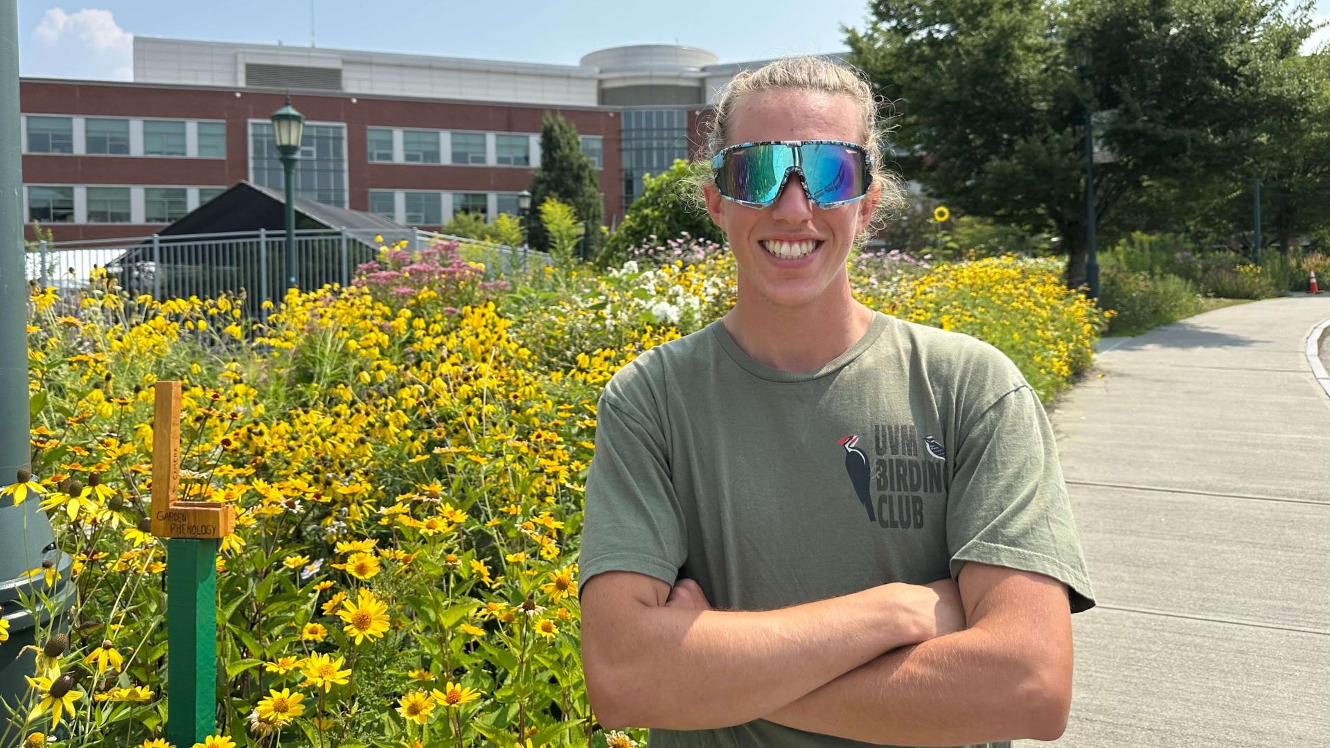 Gavin standing with his arms crossed in front of a section of the UVM gardens, with Jeffords Hall in the background.