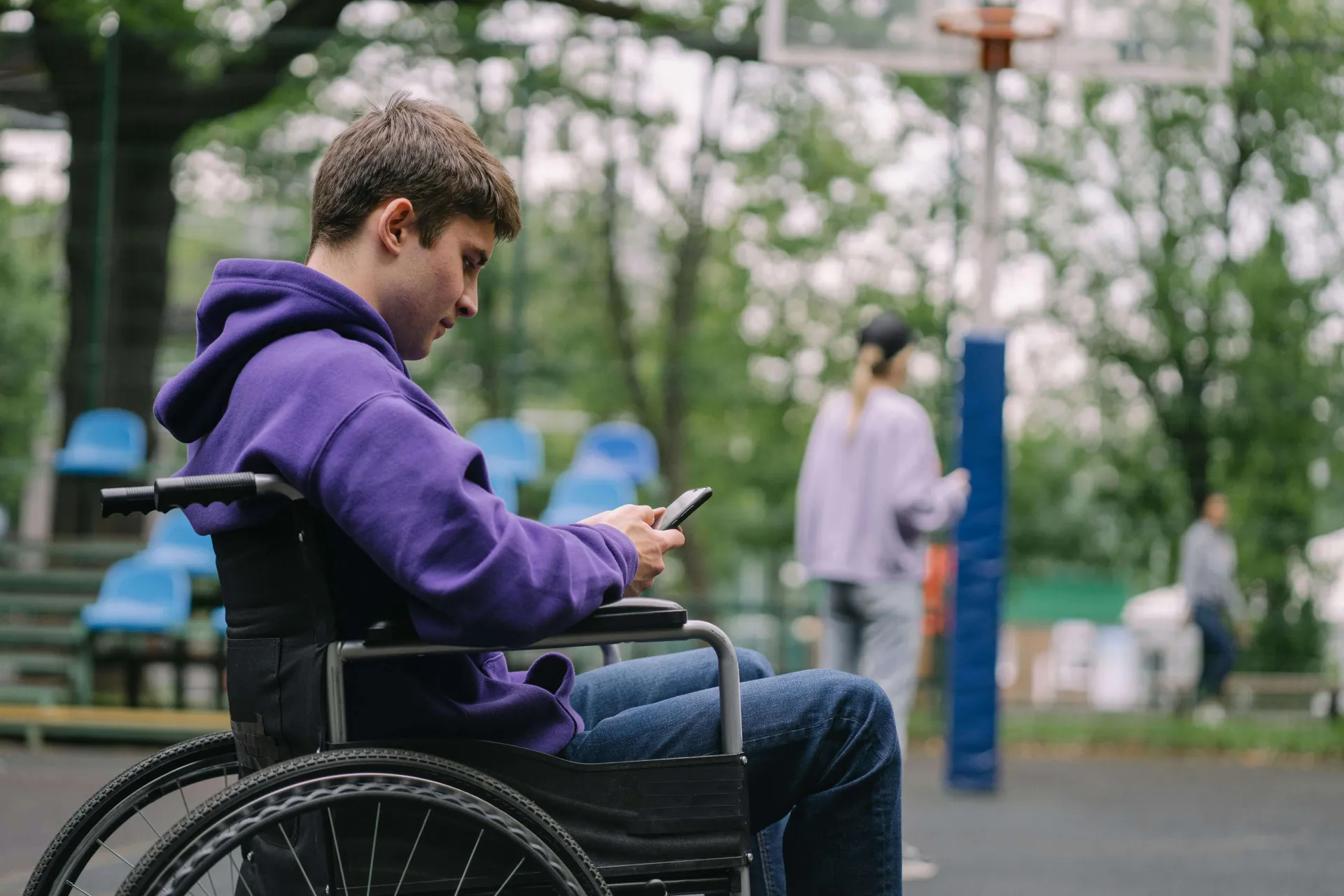 A pale-skinned college-aged boy in a deep purple sweatshirt sits in a wheelchair looking at his phone