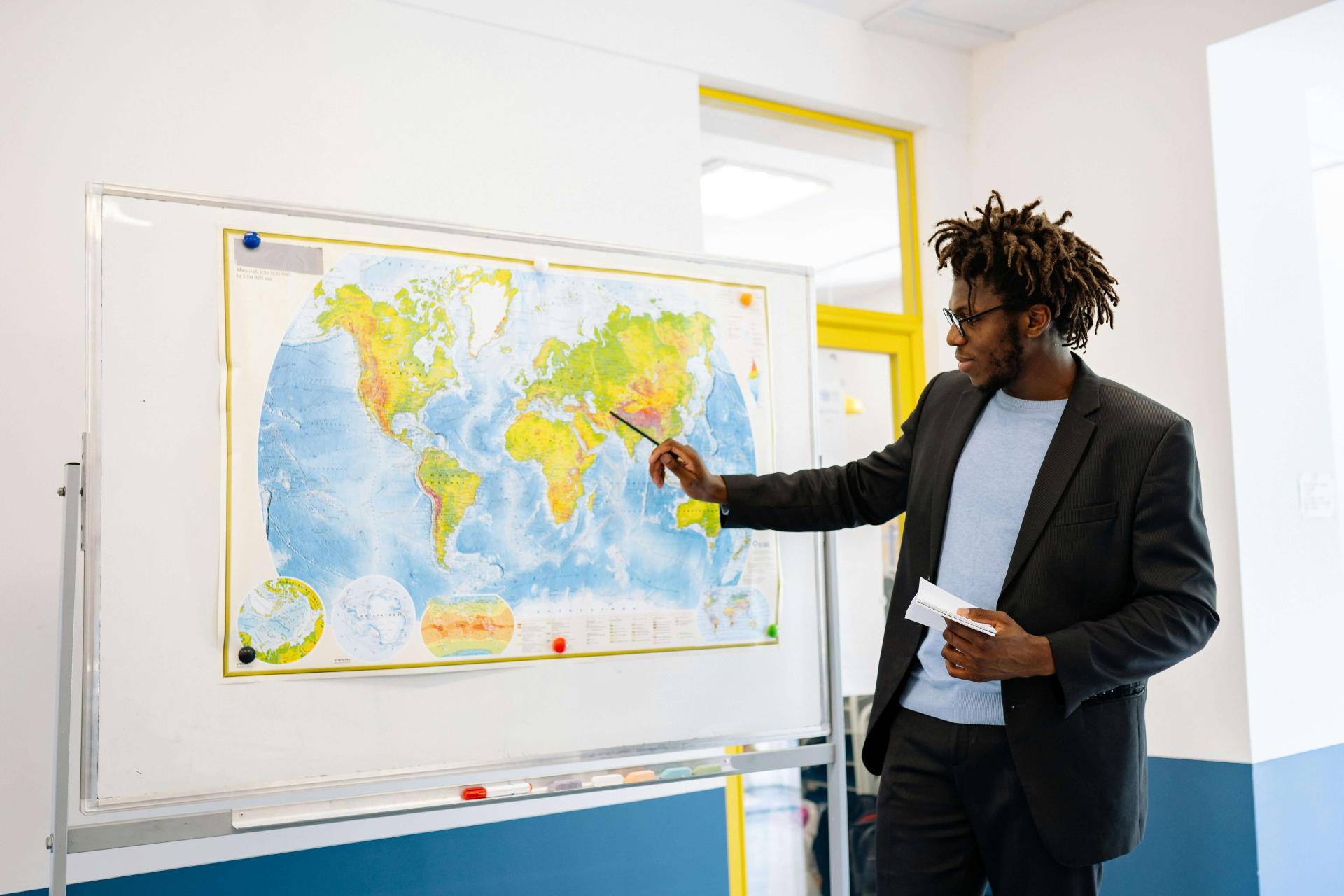 A Black man with short dreads and dark skin, dressed in a casual suit, points to a map tacked to a board in his classroom