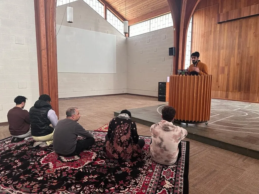 A guy wearing orange shirt is giving a sermon standing on an elevated labyrinth infront from 5 people in praying position   