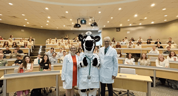 a cow mascot flanked by 2 doctors in a classroom with students