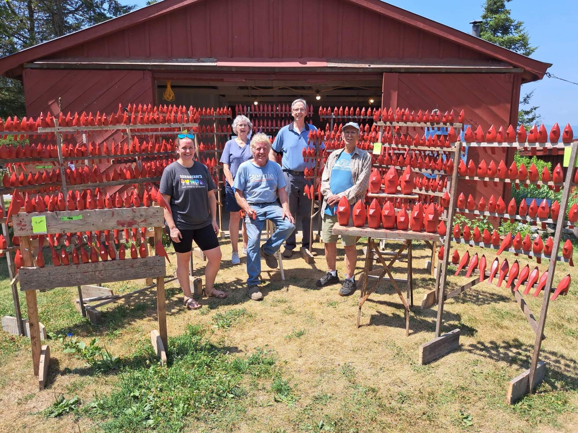 5 people in a field near a red barn surrounded by many carved wooden cardinals, also red. 