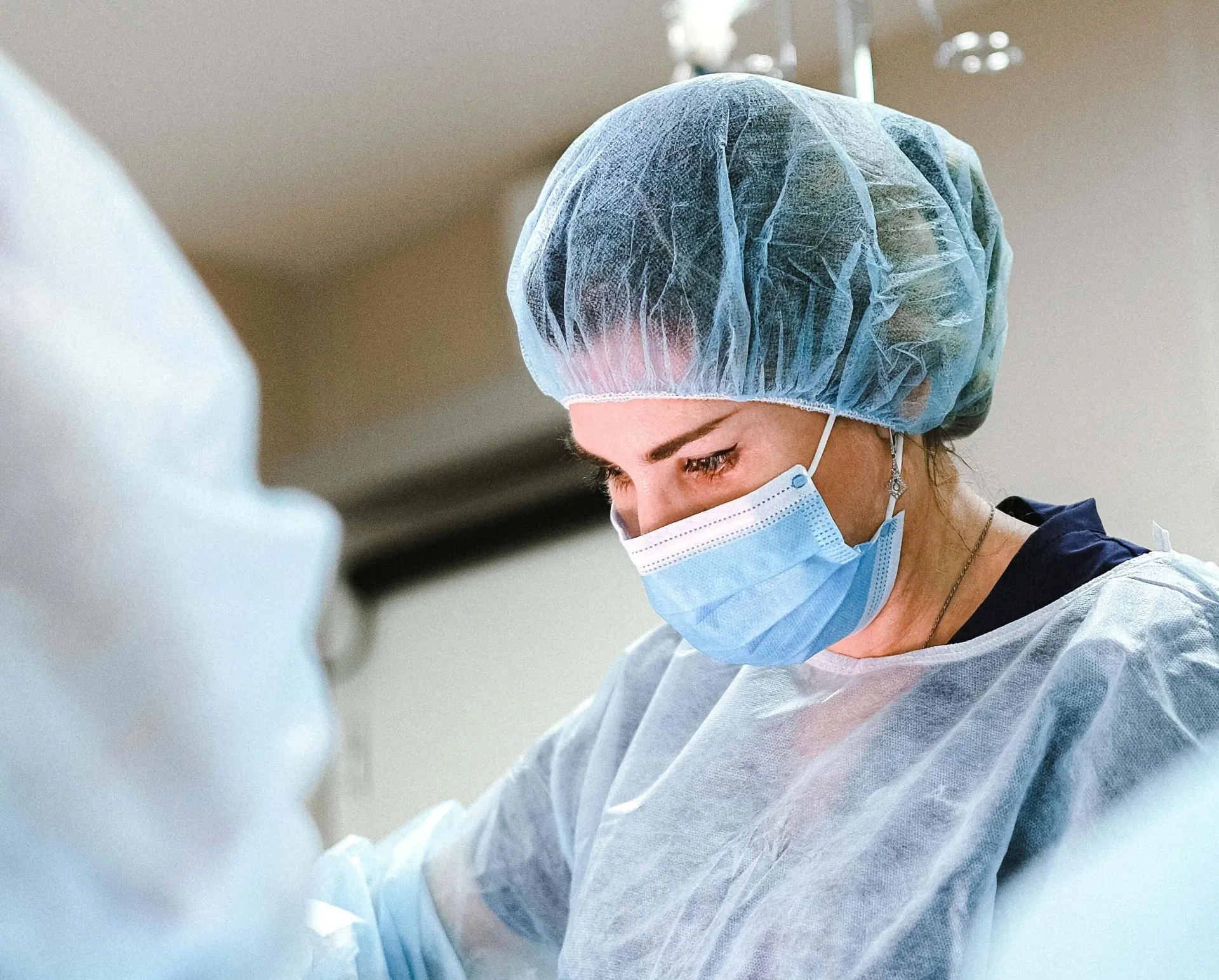 A white college-aged student in operating scrubs focuses intently on work on the operating table while a mentor looks on.