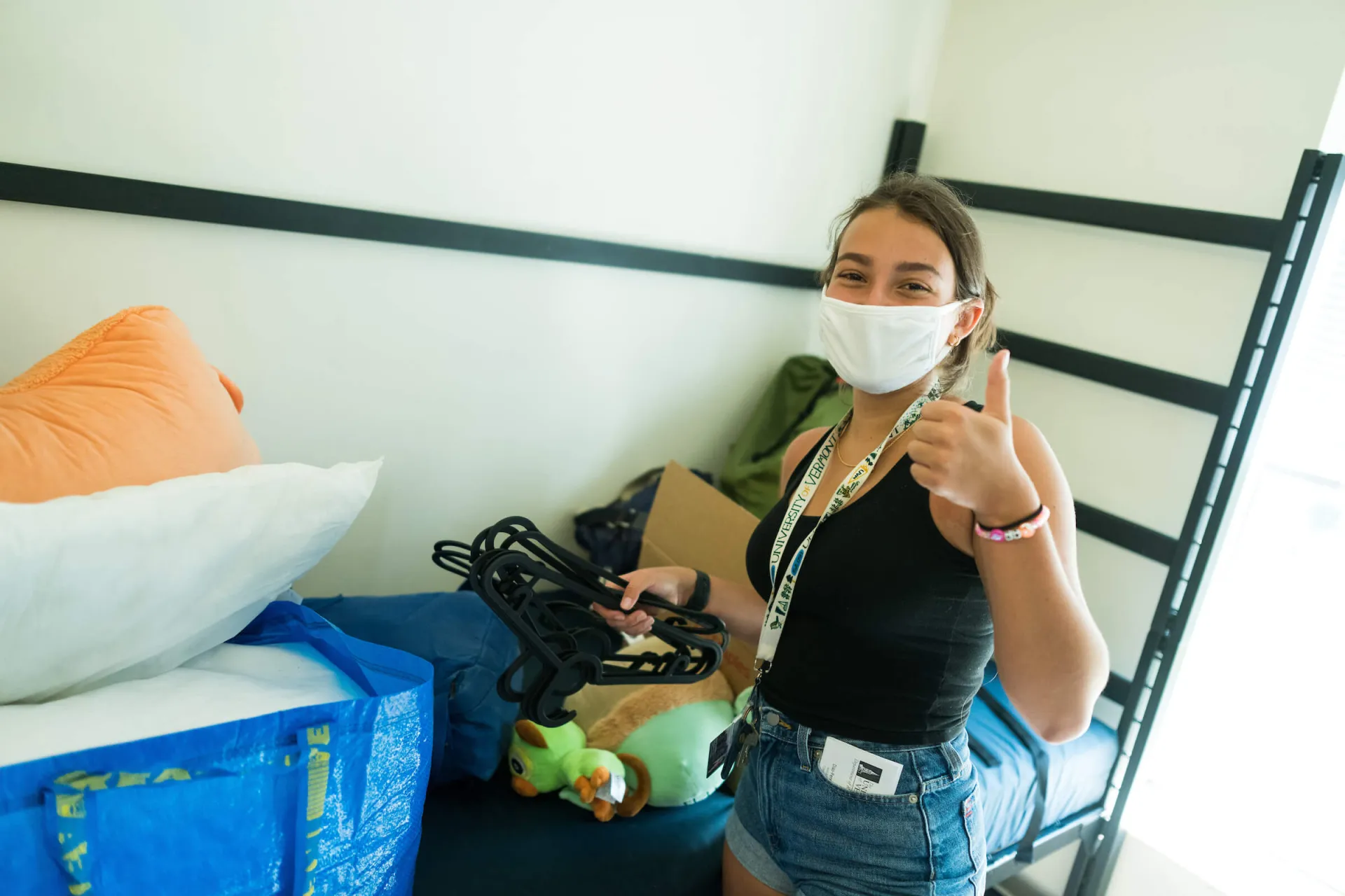 A masked, pale-skinned college-aged woman gives a thumbs-up as she unpacks her dorm