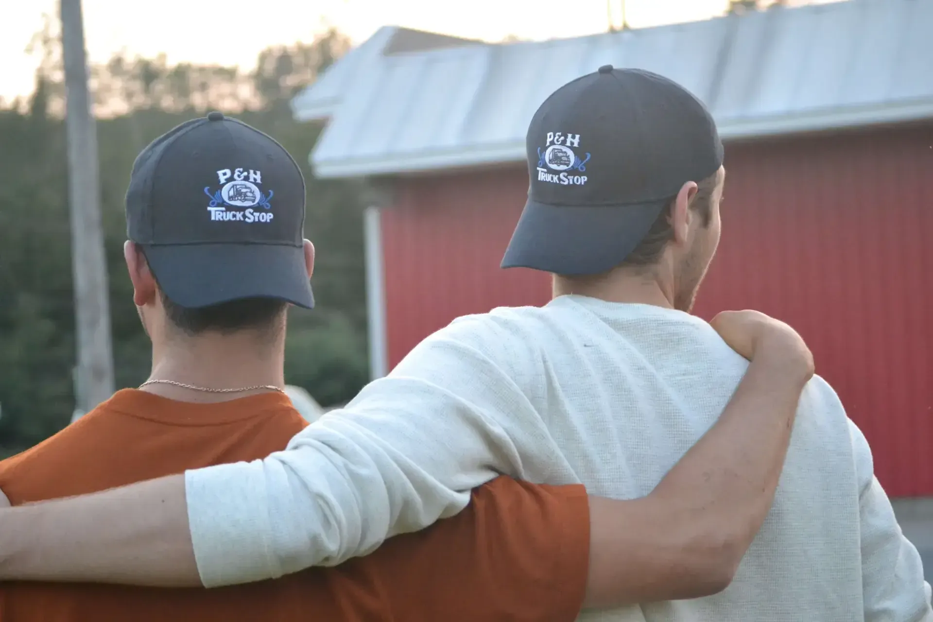 Two people stand with arms around each other, facing a red building outdoors.
