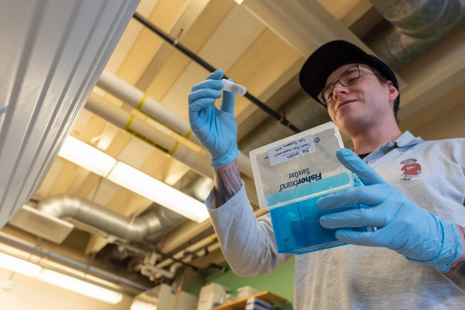 Person wearing gloves holding a small tube and a blue container in a laboratory.
