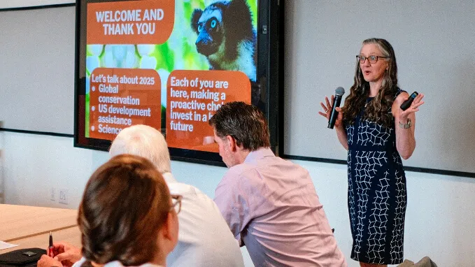 A woman holding a mic presents to people sitting at a table. A screen with a lemur on it is in the background.