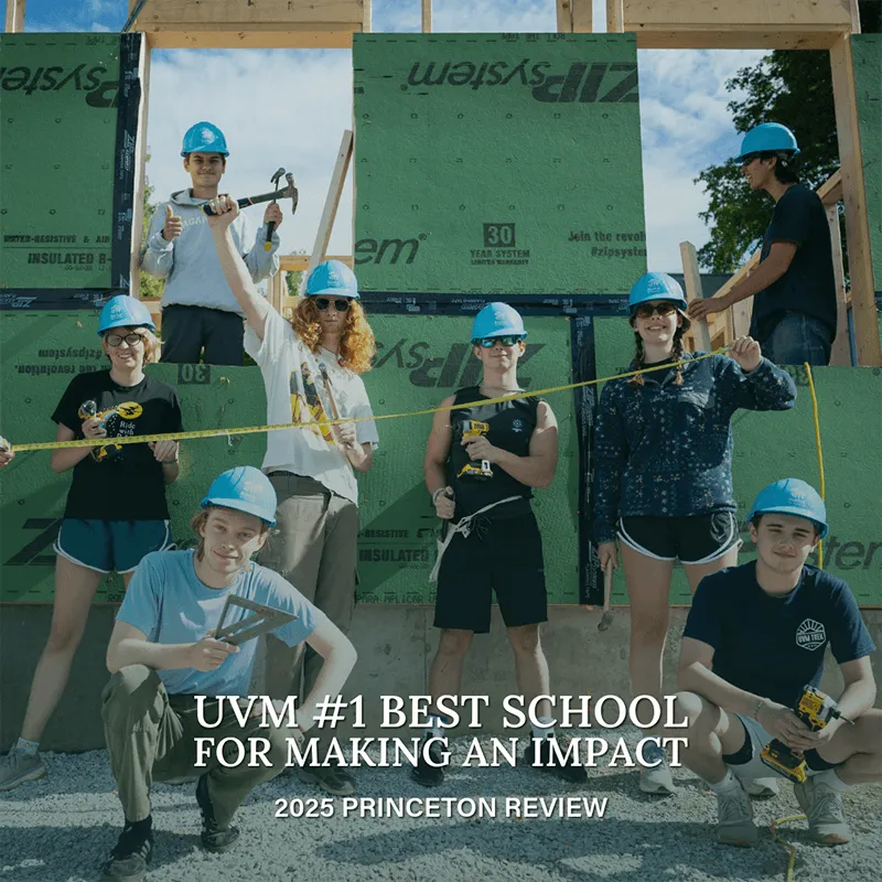Students in blue hardhats holding various tools pose in front of a Habitat for Humanity house build project.