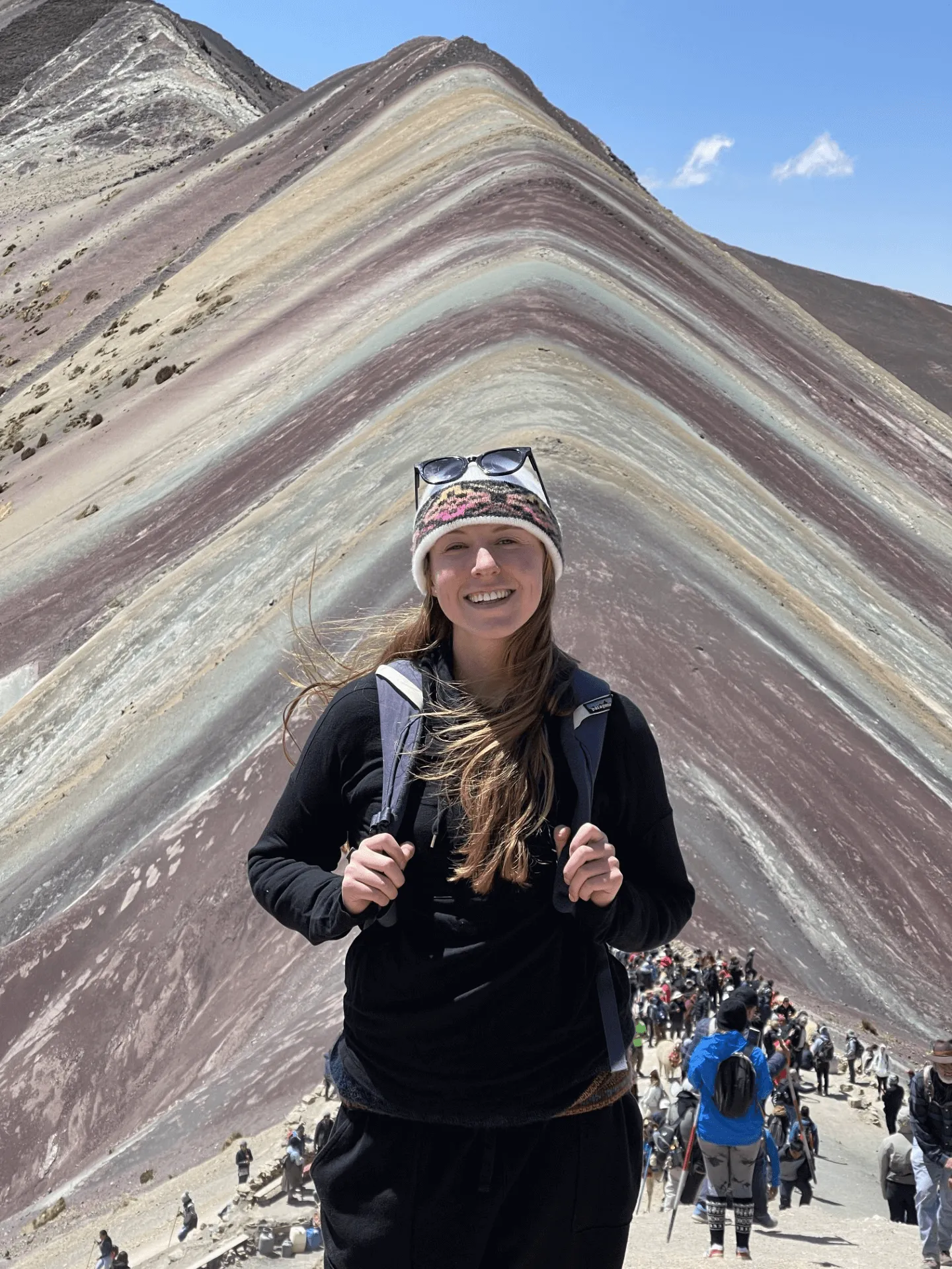 Person smiling in front of Mountain, with colorful layered slopes and hikers in the background.