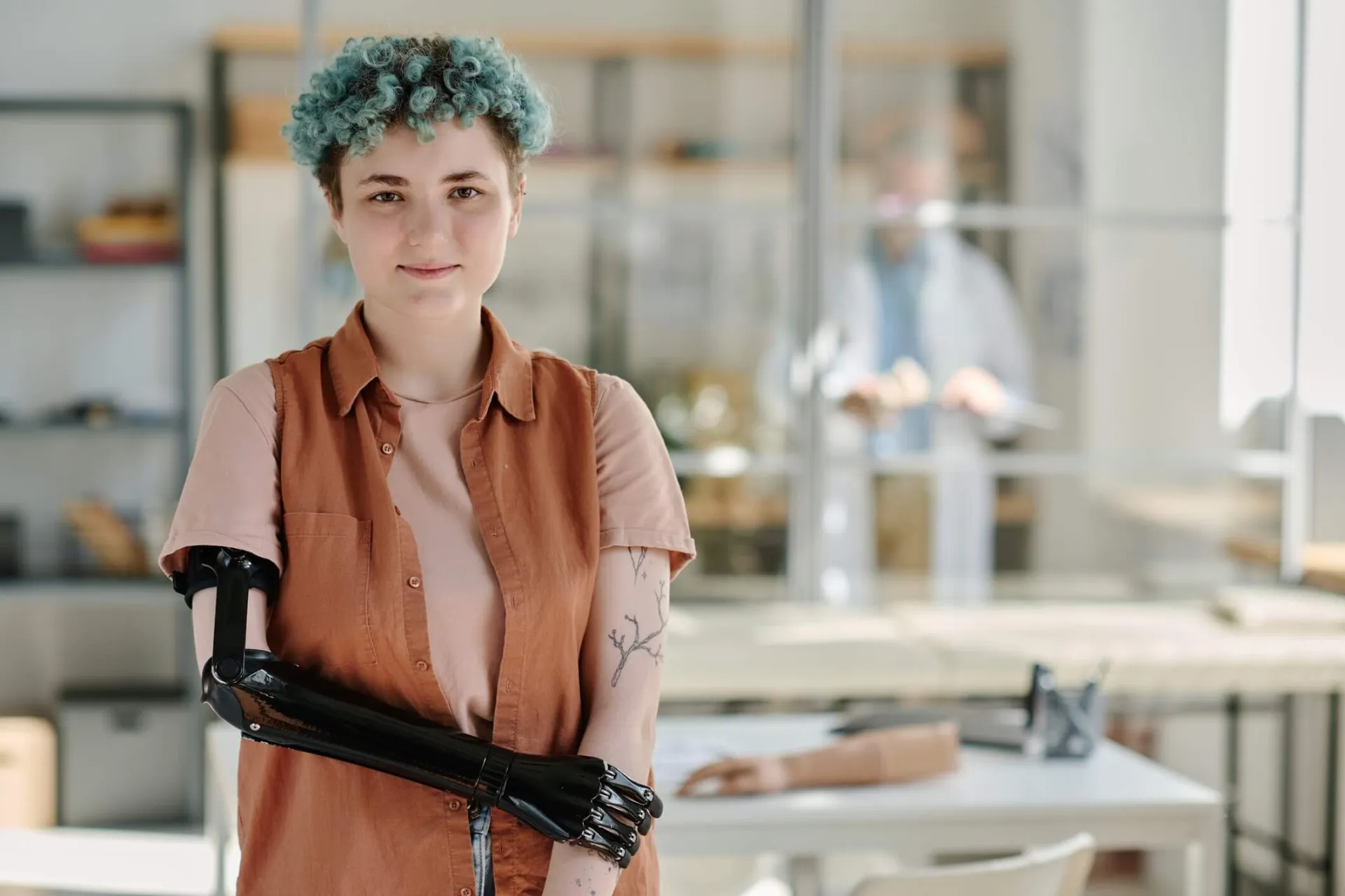 A white college-aged person with short teal curls holds their arm with a prosthetic arm crossed over their middle.