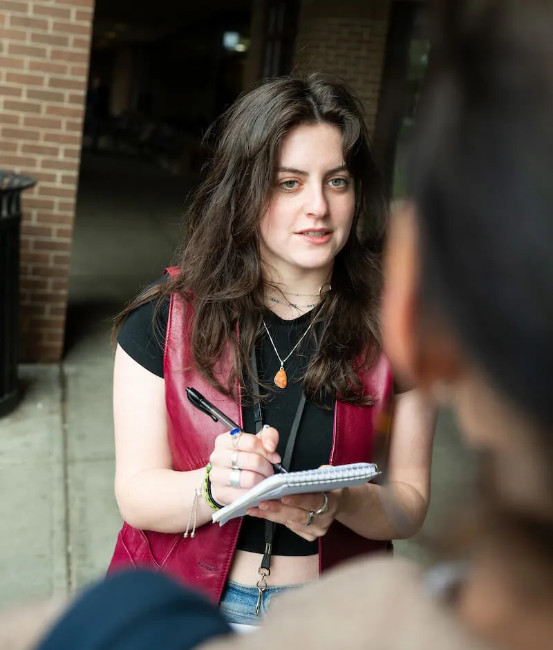 Student interviewing someone outdoors while taking notes in a small notebook.