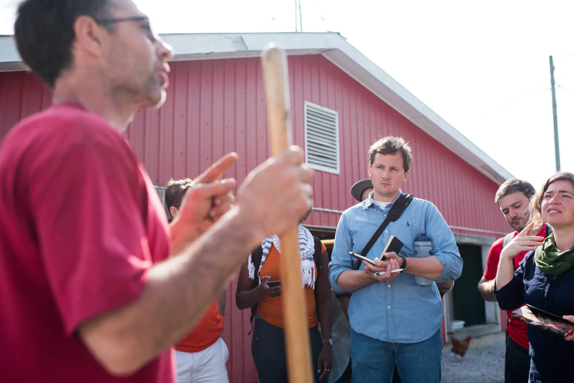 A man holds a stick and talks to a crowd in front of a barn. 
