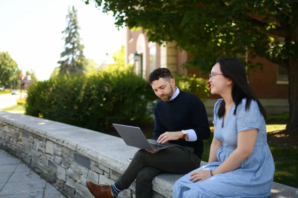 Two people sitting outside on campus, one using a laptop during a conversation.