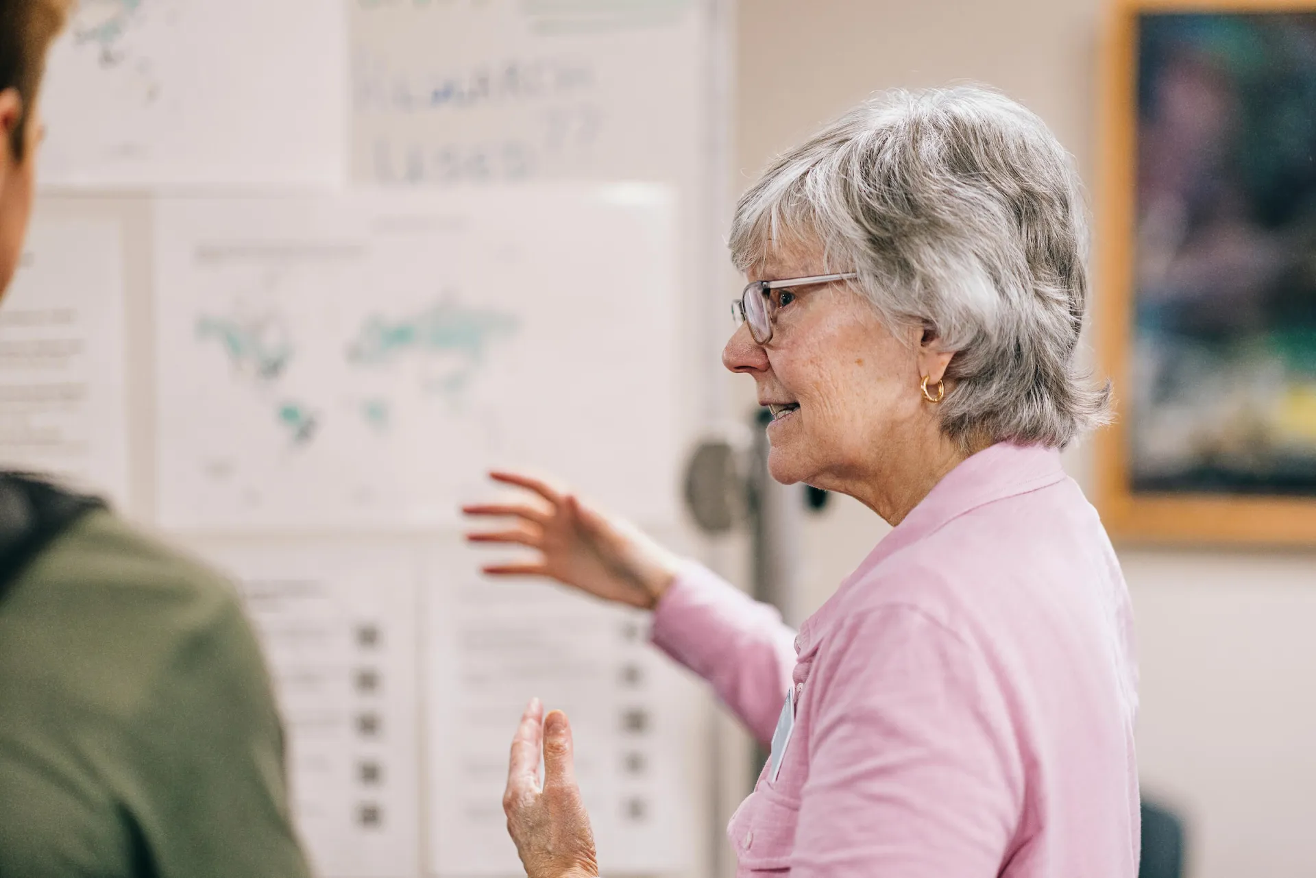 woman gesturing toward materials on the wall behind her as she speaks