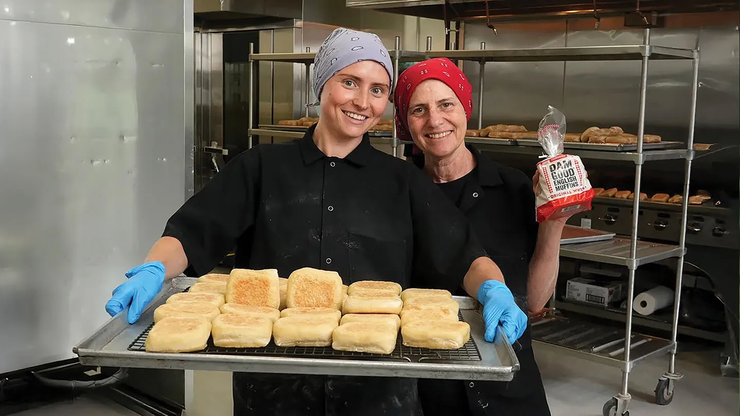 two people stand in an industrial kitchen holding loaves of bread