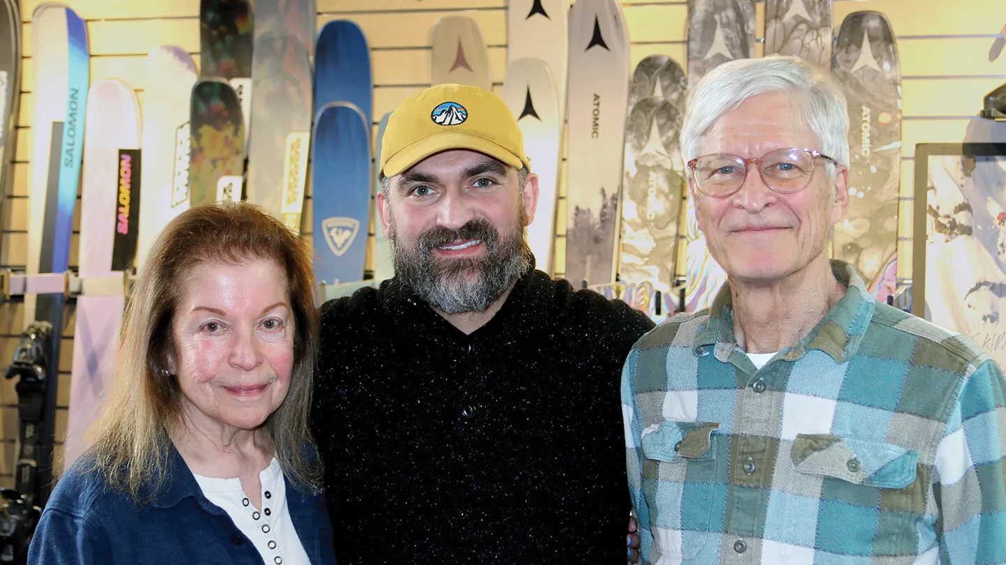 three people smiling at the camera in front of a rack of skis