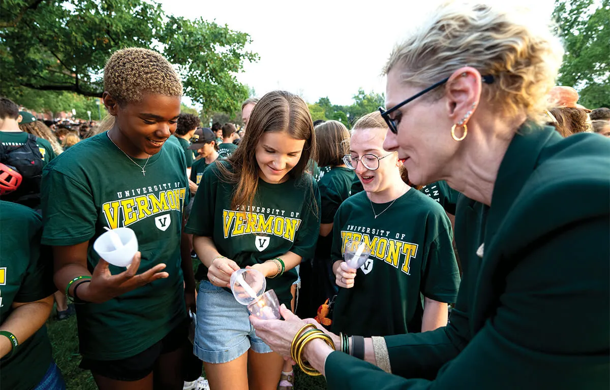 marlene tromp lights a candle with three students wearing green shirts with the text Vermont on them