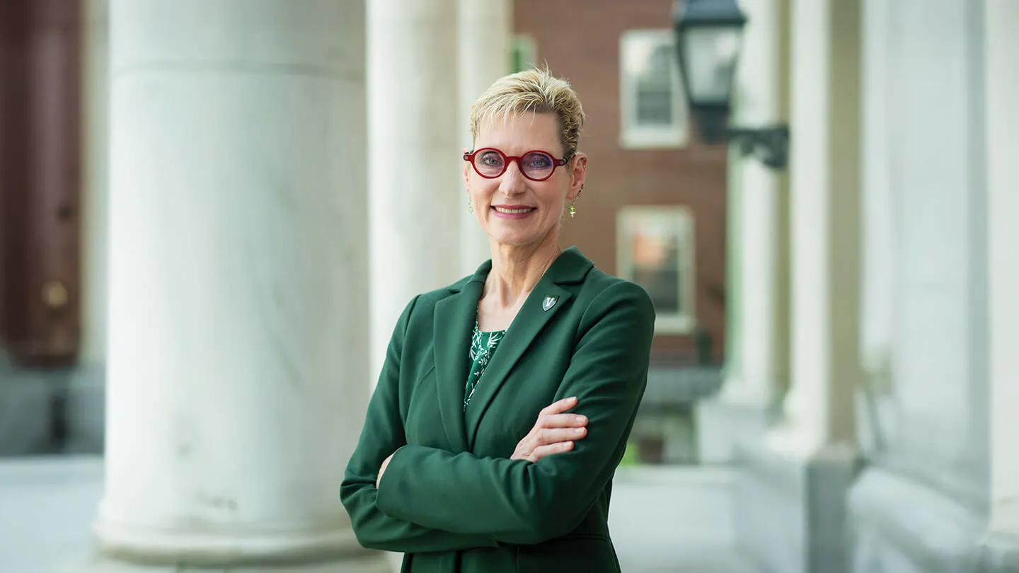 marlene tromp poses with arms crossed in a green blazer and red glasses in front of white columns