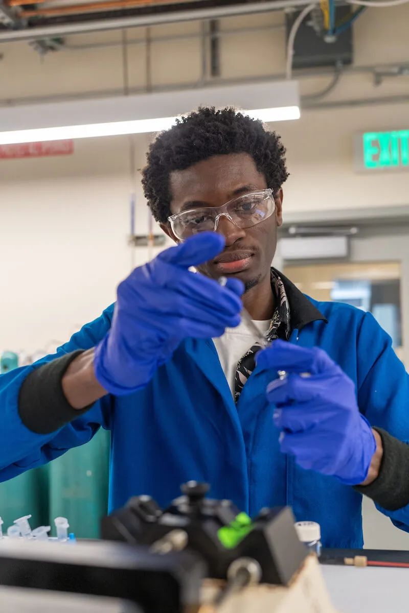 Student in a lab coat and gloves handling equipment while wearing safety goggles.
